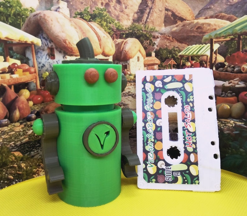 May include: A green robot figure with brown accents and a circular emblem. Beside it is a cassette tape labeled "Vegan Robot Food" and "Hot Salsa" with a colorful vegetable pattern. The robot has a plant-based symbol.