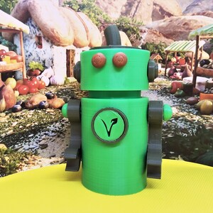 May include: A green robot figurine with brown accents, including eyes, arms, and a top hat. A circular emblem with a leaf design is on the robot's chest. The robot is on a yellow surface with a blurred background of a market scene.