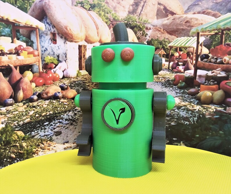 May include: A green robot figurine with brown accents, including eyes, arms, and a top hat. A circular emblem with a leaf design is on the robot's chest. The robot is on a yellow surface with a blurred background of a market scene.