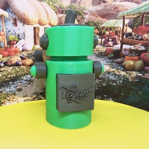 May include: A green and gray 3D-printed robot with the text "Vegan Life" on its chest. The robot has a cylindrical body, a boxy head, and small arms. The background features a colorful market scene.