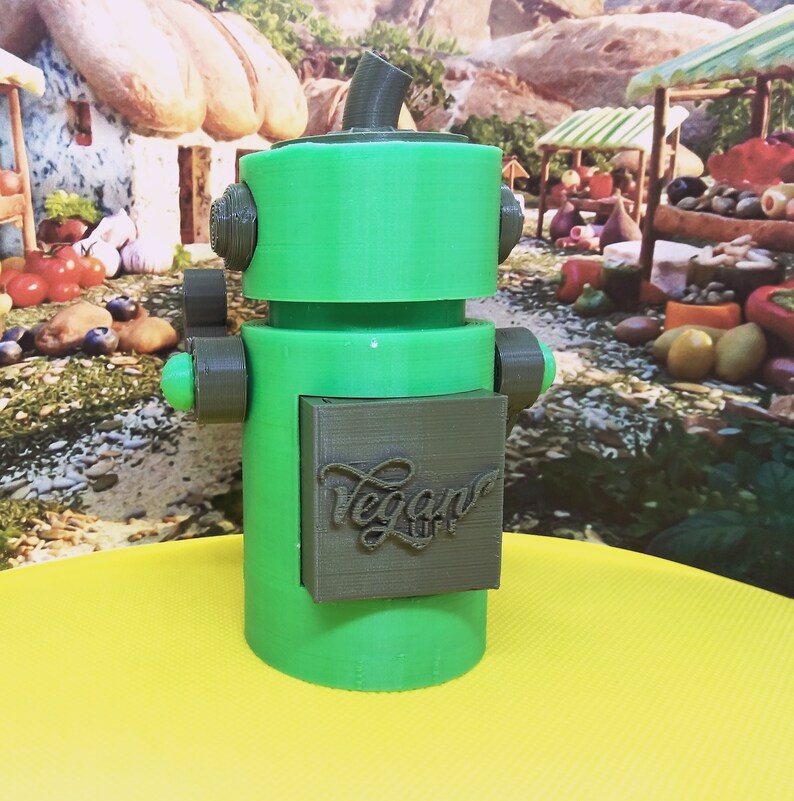 May include: A green and gray 3D-printed robot with the text "Vegan Life" on its chest. The robot has a cylindrical body, a boxy head, and small arms. The background features a colorful market scene.