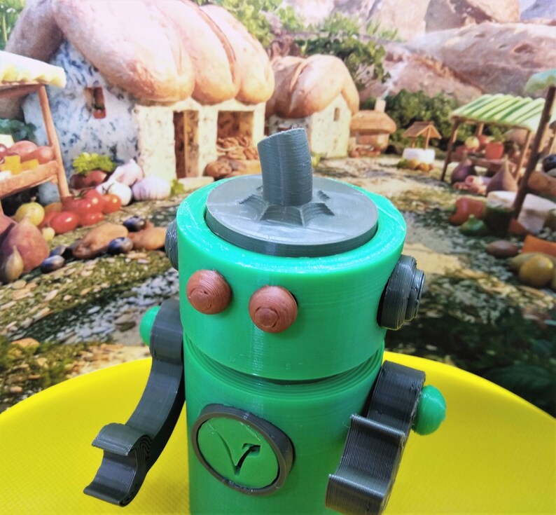 May include: A green robot toy with brown circular eyes, a gray top, and gray arms. The robot has a circular emblem with a V-shaped design. The background features a whimsical village scene with miniature buildings and market stalls.