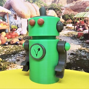 May include: A green robot figurine with brown accents. The robot has round eyes, a top hat, and a circular emblem with a leaf design. The arms and legs are articulated. The robot is standing on a yellow surface.