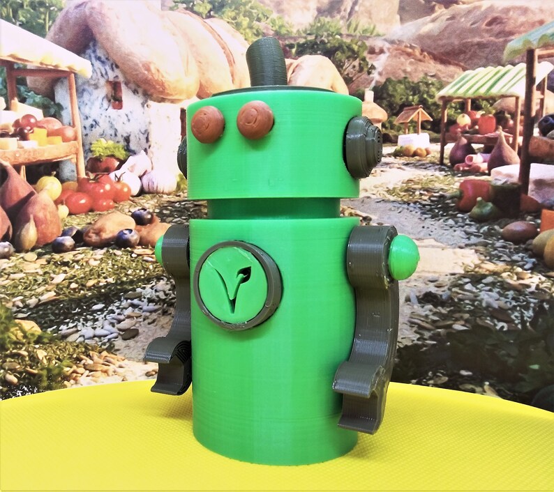 May include: A green robot figurine with brown accents. The robot has round eyes, a top hat, and a circular emblem with a leaf design. The arms and legs are articulated. The robot is standing on a yellow surface.