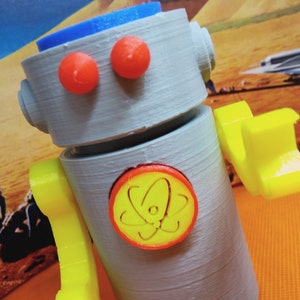 May include: A gray robot toy with orange eyes, yellow arms, and a blue head. The robot has a yellow and red emblem on its chest. The robot is a fun, colorful toy.