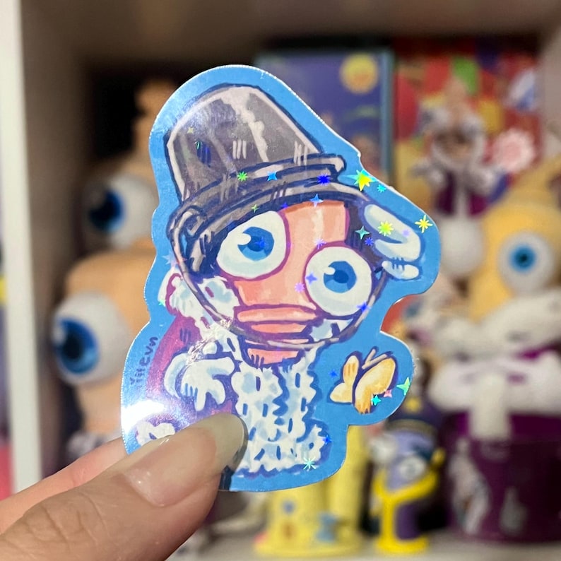 Pode incluir: Um autocolante azul com uma personagem de desenho animado com grandes olhos azuis, um chap&eacute;u alto e uma roupa branca. O autocolante tem um efeito hologr&aacute;fico com detalhes brilhantes. A personagem segura um pequeno objeto amarelo.