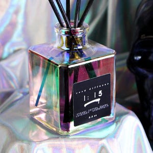 May include: A square glass diffuser bottle with a holographic finish, containing black reeds. The bottle has a black label with white text that reads "1:15". A black skull is in the background. The product is a home fragrance.