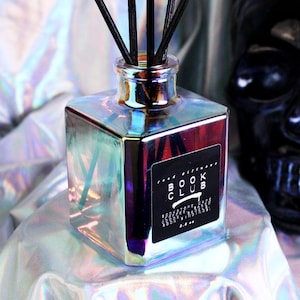 Puede incluir: Botella difusora de vidrio iridiscente con varillas negras. La botella cuadrada tiene una etiqueta con texto y está sobre un fondo holográfico. Una calavera negra está en el fondo. Producto para fragancia del hogar.