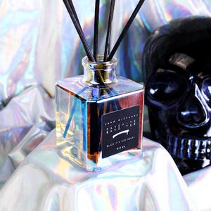 May include: A clear, square glass diffuser bottle with black reeds. The label reads "Favorite Band Tee, Black Fig and Honey." A black skull is in the background, and the background is a shiny, iridescent fabric.