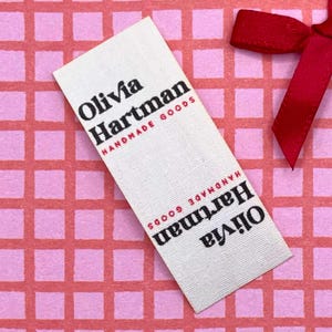 Può includere: Un'etichetta di tessuto bianco con la scritta "Olivia Hartman HANDMADE GOODS" in nero e rosso. L'etichetta è su uno sfondo a griglia rosa con un nastro rosso.