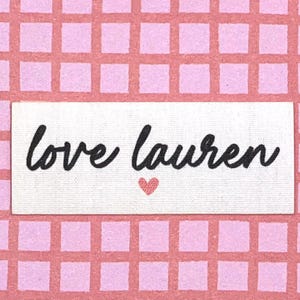 May include: A white rectangular label with the words "love lauren" in black script, and a small pink heart. The label is set against a pink grid background.