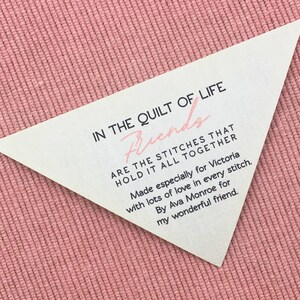 Könnte beinhalten: Ein dreieckiges Etikett mit Text auf einem rosa strukturierten Hintergrund. Das Etikett lautet "IN THE QUILT OF LIFE Friends ARE THE STITCHES THAT HOLD IT ALL TOGETHER." Es enthält auch eine Widmung an Victoria.
