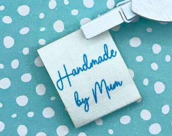 Op de afbeelding: Een klein, rechthoekig wit stoffen label met de woorden "Handmade by Mum" in blauw script. Het label wordt vastgehouden door een witte clip tegen een lichtblauwe achtergrond met witte stippen. Het label is voor handgemaakte ambachten.