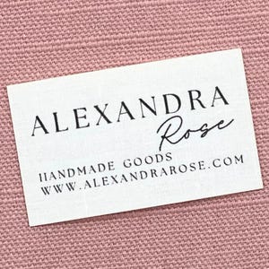 Può includere: Un'etichetta rettangolare bianca con testo nero. Il testo recita "ALEXANDRA Rose" in una combinazione di caratteri script e sans-serif. Sotto, si legge "HANDMADE GOODS" e "WWW.ALEXANDRAROSE.COM". Lo sfondo è in tessuto rosa intrecciato.