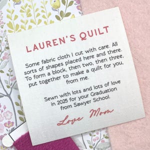 Può includere: Un'etichetta di tessuto bianco con la scritta "LAUREN'S QUILT" in rosso. L'etichetta include testo scritto a mano sulla creazione della trapunta e un messaggio d'amore da "Mamma". L'etichetta è posta su uno sfondo a fantasia floreale.