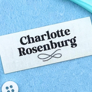 May include: A white rectangular label with the text "Charlotte Rosenburg" in black, with a decorative infinity symbol below the name. The label is on a light blue textured surface, with a white button and a turquoise object in the background.