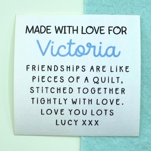 Può includere: Un'etichetta di tessuto bianco con testo nero e blu. Il testo recita: "MADE WITH LOVE FOR Victoria. FRIENDSHIPS ARE LIKE PIECES OF A QUILT, STITCHED TOGETHER TIGHTLY WITH LOVE. LOVE YOU LOTS LUCY XXX."