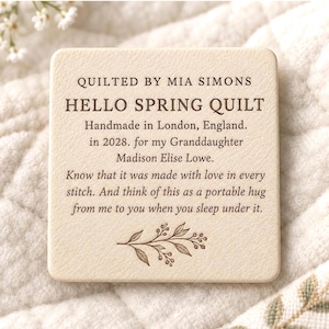 Puede incluir: Una etiqueta cuadrada de color crema con textura y esquinas redondeadas que presenta un texto impreso en marrón oscuro: QUILTED BY MIA SIMONS, HELLO SPRING QUILT, Handmade in London, England, in 2028, for my Granddaughter Madison Elise Lowe.