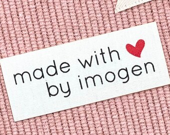 May include: A white rectangular tag with the text "made with by imogen" in black, and a red heart. The tag is on a pink textured fabric background, with a white ribbon in the upper right corner.