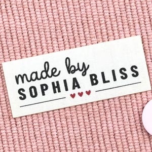 May include: A rectangular label with the text "made by SOPHIA BLISS" in black script and block letters. Three small red hearts are below the text, and the label is on a pink textured surface.