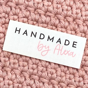May include: A white rectangular label with the words "HANDMADE" in black capital letters and "by Alexa" in pink cursive script. The label is set against a background of a pink crocheted fabric.
