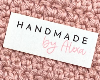 May include: A white rectangular label with the words "HANDMADE" in black capital letters and "by Alexa" in pink cursive script. The label is set against a background of a pink crocheted fabric.