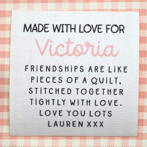 Può includere: Un'etichetta di tessuto bianco con testo nero che recita "MADE WITH LOVE FOR Victoria". Sotto, c'è scritto "FRIENDSHIPS ARE LIKE PIECES OF A QUILT, STITCHED TOGETHER TIGHTLY WITH LOVE. LOVE YOU LOTS LAUREN XXX."
