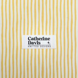 May include: A fabric with a repeating pattern of vertical yellow stripes on a white background. A rectangular label with the text "Catherine Davis KNITTED DESIGNS" is centered on the fabric.
