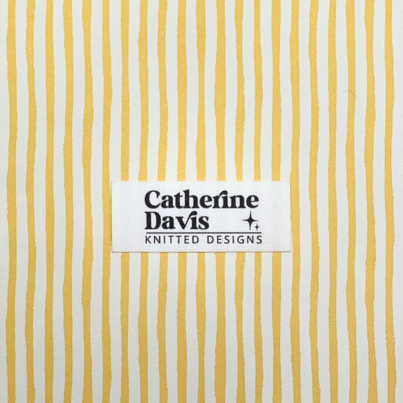 May include: A fabric with a repeating pattern of vertical yellow stripes on a white background. A rectangular label with the text "Catherine Davis KNITTED DESIGNS" is centered on the fabric.