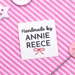 Könnte beinhalten: Ein quadratisches, cremefarbenes Stoffetikett mit dem Text "Handmade by ANNIE REECE" in Schwarz und einer roten Schleife. Das Etikett befindet sich auf einem rosa-weiß gestreiften Hintergrund, mit weißen und rosa Knöpfen.