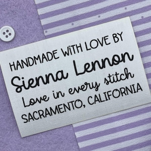May include: A white rectangular label with black text that reads "HANDMADE WITH LOVE BY Sienna Lennon Love in every stitch SACRAMENTO, CALIFORNIA." The label is on a purple background with a white button.