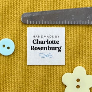 Puede incluir: Una etiqueta cuadrada de tela blanca con el texto "HANDMADE BY Charlotte Rosenburg" en negro y un símbolo de infinito azul claro. También se ven una regla, un botón azul claro y un botón amarillo en forma de flor.
