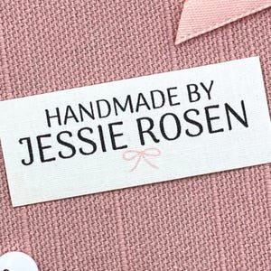 May include: A white rectangular label with the text "HANDMADE BY JESSIE ROSEN" in black, with a small pink bow graphic. The label is on a pink woven fabric background, with a pink ribbon and a white button visible.