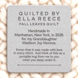 May include: A square light gray leather label with rounded corners featuring dark gray text that reads: QUILTED BY ELLA REECE FALL LEAVES QUILT. Handmade in Manhattan, New York, in 2028. for my Granddaughter Madison Joy Monroe. Know that it was made with love in every stitch. And think of this as a portable hug from me to you when you sleep under it.