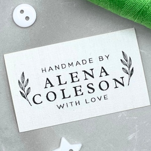 May include: A rectangular white fabric label with the text "HANDMADE BY ALENA COLES ON WITH LOVE" in black. The label is decorated with leaf sprigs and is surrounded by a white button and green thread.