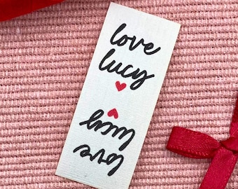 May include: A white rectangular label with the words "love lucy" in black script, repeated with a red heart between each word. The label is on a pink textured surface, with red ribbon and thread in the background.