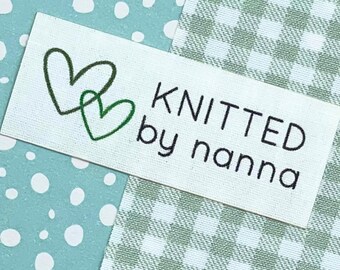 May include: A rectangular white label with the text "KNITTED by nanna" in black. Two intertwined green heart outlines are in the upper left corner. The label is on a background of light blue polka dots and green gingham.