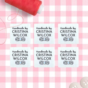 May include: Six white fabric labels with the text "Handmade by CRISTINA WILCOX" and a spool of thread graphic. A red spool of thread and a pink gingham background are also visible.