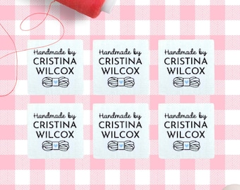 Op de afbeelding: Zes witte stoffen labels met de tekst "Handmade by CRISTINA WILCOX" en een garenspoelafbeelding. Een rode garenspoel en een roze ruitjesachtergrond zijn ook zichtbaar.