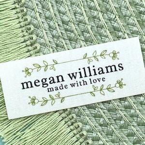 May include: A rectangular white label with the text "megan williams made with love" in black. The label is framed with green floral designs and rests on a textured green surface with fringe.
