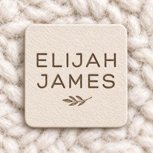 Könnte beinhalten: Ein quadratisches, cremefarbenes Kunstlederetikett mit abgerundeten Ecken. Der Text ELIJAH JAMES ist mittig in einer dunkelbraunen, serifenlosen Schrift eingeprägt, mit einer kleinen, detaillierten Abbildung eines Blattzweigs unter dem Namen.