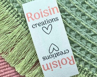 May include: A white fabric label with the text "Roisin creations" in coral-colored letters, along with two black heart symbols. The label is set against a textured background with green and pink hues.
