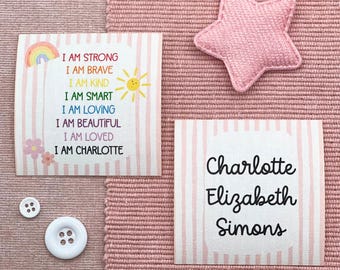 Op de afbeelding: Twee wit en roze gestreepte stoffen labels met tekst, een roze ster-vormige stoffen patch en twee witte knopen. Eén label leest "I AM STRONG" en "I AM CHARLOTTE". Het andere label leest "Charlotte Elizabeth Simons".