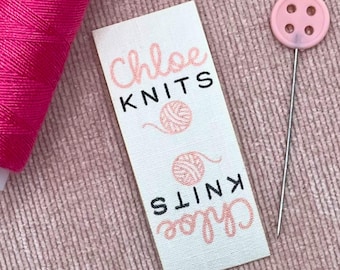 Op de afbeelding: Een wit stoffen label met de tekst "Chloe KNITS" in roze en zwart, met illustraties van garenballen. Een roze knoop met vier gaten en een lange speld zijn ook zichtbaar, samen met een spoel roze garen. De items liggen op een roze, getextureerd oppervlak.