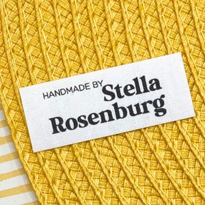 May include: A white rectangular label with the text "HANDMADE BY Stella Rosenburg" in black lettering. The label is placed on a textured yellow surface with a woven pattern. The background has a yellow and white striped pattern.