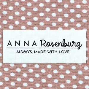May include: A white rectangular label with the text "ANNA Rosenburg" in black script, with the words "ALWAYS, MADE WITH LOVE" below. The label is set against a background of pink fabric with white polka dots.