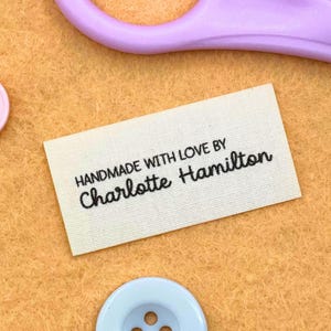 May include: A rectangular, off-white fabric label with the text "HANDMADE WITH LOVE BY Charlotte Hamilton" in black script. The label is positioned on a textured, light orange surface, with a pair of purple scissors and two buttons in the background.