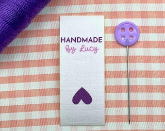 Op de afbeelding: Een wit stoffen label met de tekst "HANDMADE by Lucy" in paars script, en een paars hart. Een paarse knoop met een speld en een spoel paars garen zijn ook zichtbaar. De achtergrond is een roze en wit geruit patroon.