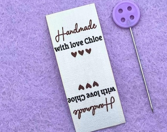 May include: A rectangular, off-white fabric tag with the words "Handmade with love Chloe" in brown script, along with three heart symbols. A purple button with four holes is attached to a silver pin. The background is a light purple fabric.