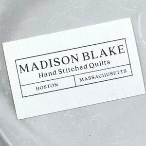 May include: A white rectangular label with black text. The label reads "MADISON BLAKE" at the top, "Hand Stitched Quilts" in the middle, and "BOSTON" and "MASSACHUSETTS" at the bottom, all within a black border.